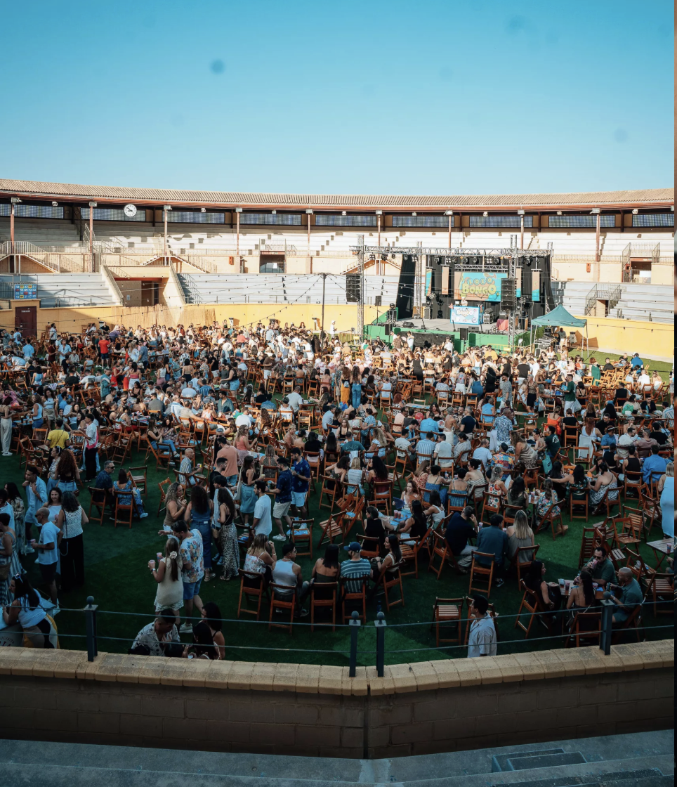 Vista desde las gradas de la Plaza de Toros de Torremolinos llena de público durante Loco Bongo en Plaza Paraíso: el ruedo con césped, sillas de madera, el escenario al fondo y cientos de personas disfrutando al aire libre